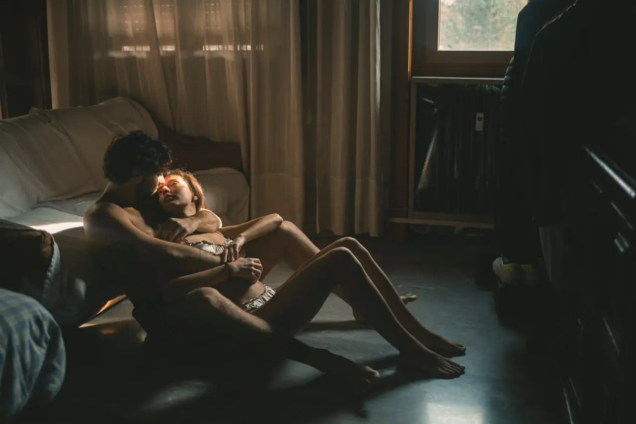 Un couple assis au sol, enlacé et s’embrassant, illustrant la complicité, l’intimité et le plaisir partagé à deux.