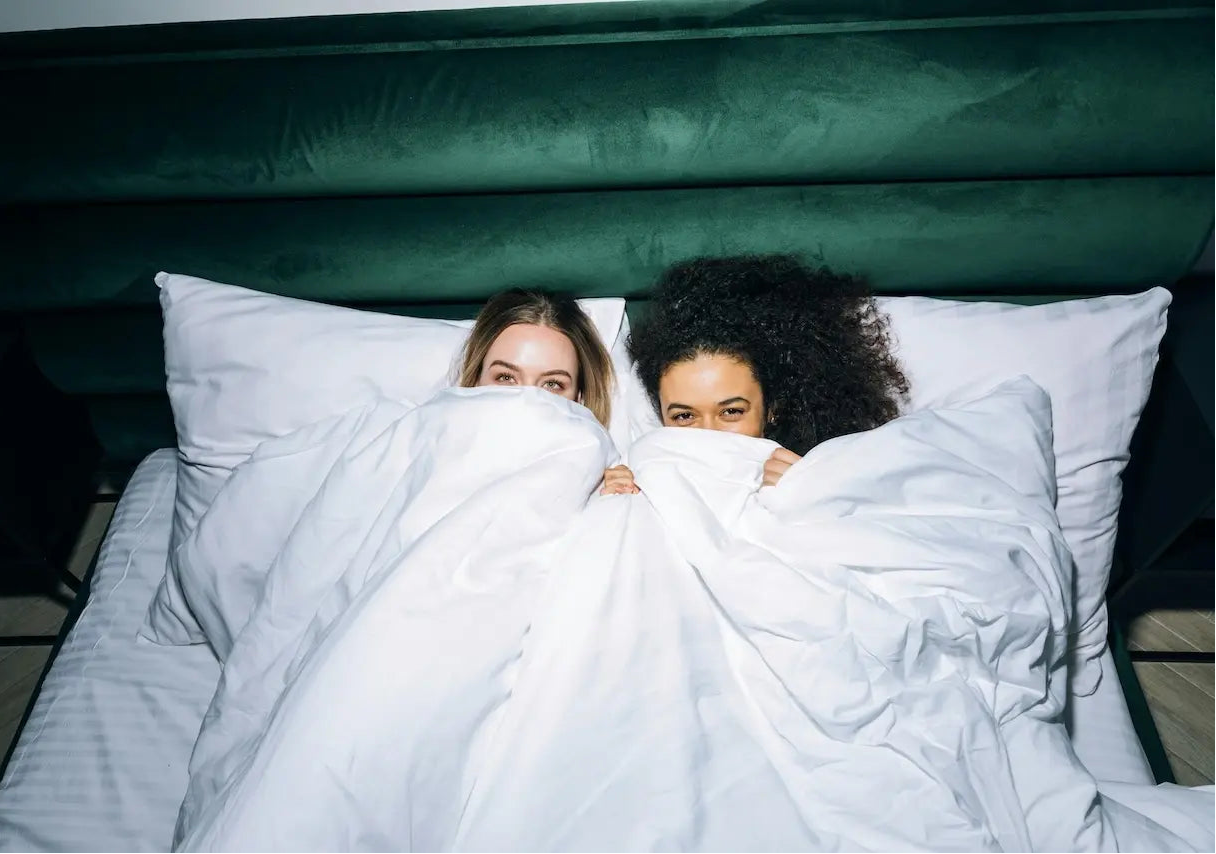 Deux femmes allongées dans un lit, la couette tirée devant leur visage